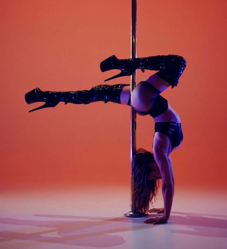 Dance instructor posing in a handstand against a pole in vivid orange lighting